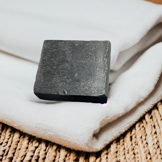  A square black soap bar resting on a stack of folded white towels, with a woven surface underneath, suggesting a minimalist and clean skincare product.