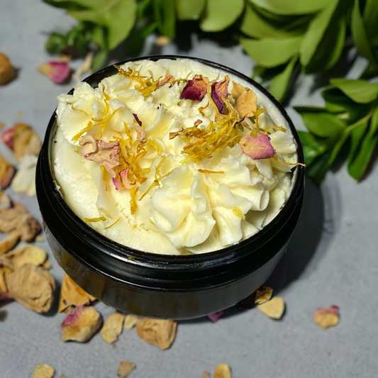  A black jar of Nature's Butter Balm scented with vanilla oil is displayed, featuring a rich, creamy swirled texture. The top is adorned with delicate dried flower petals, adding a natural, artisanal touch. The jar sits against a soft gray background, surrounded by scattered petals, evoking a sense of handcrafted luxury and botanical goodness. Lush green leaves in the background further emphasize the balm's natural ingredients and nourishing properties. Handcrafted in Dallas, Arlington, Ft. Worth TX.