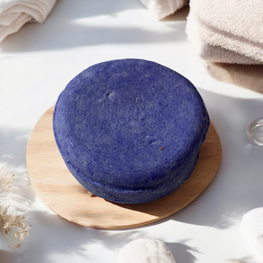 : A round purple shampoo bar placed on a wooden tray, surrounded by soft towels and natural elements, suggesting a clean and refreshing haircare product. Handcrafted in Dallas, Arlington, Ft. Worth, Grand Prairie, Midlothian, TX.
