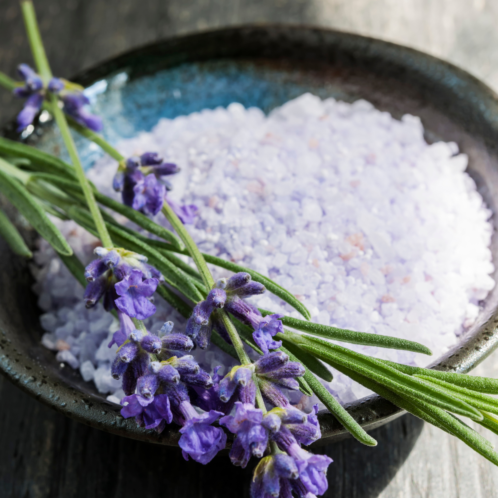 A black ceramic bowl filled with light purple lavender soaking salts, topped with sprigs of lavender flowers and stems, set against a rustic wooden background. 